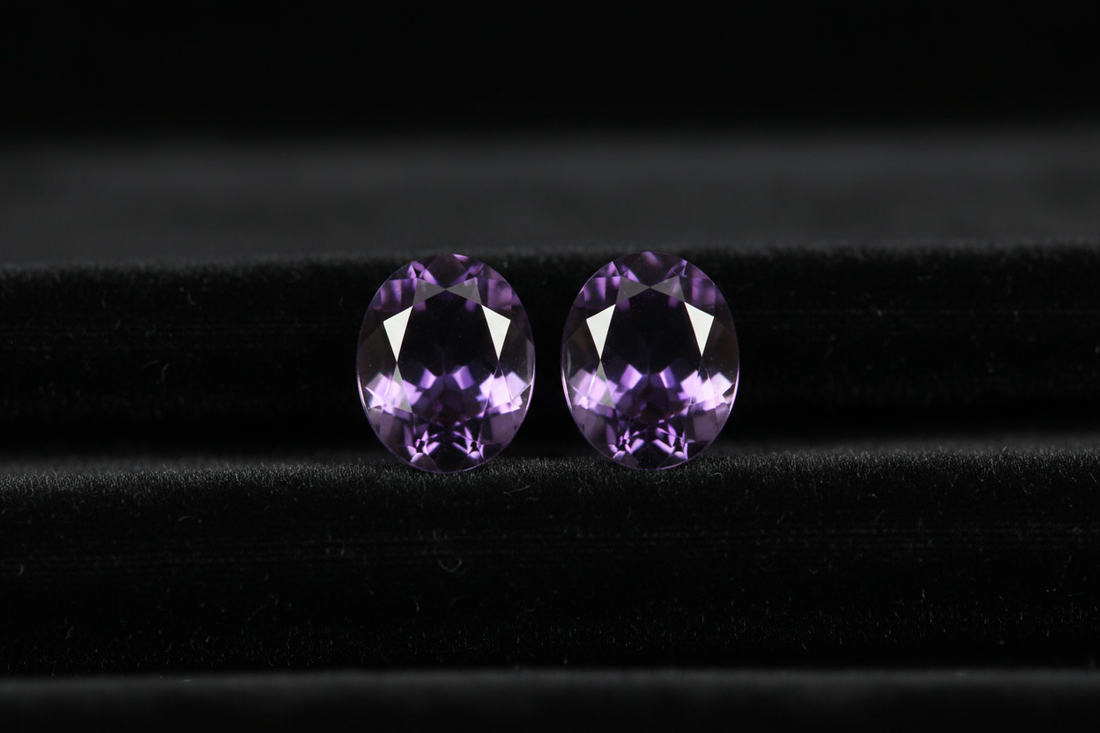Perfectly matched purple amethyst gemstone pair on black velvet - identical 2.5ct oval cut stones with same deep royal purple color and eye-clean clarity demonstrating why matched pairs cost 3X more and take 6 months to find